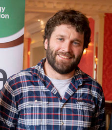 Dr Maurice Deasy, smiling, wearing a blue checked shirt, standing in front of a conference banner